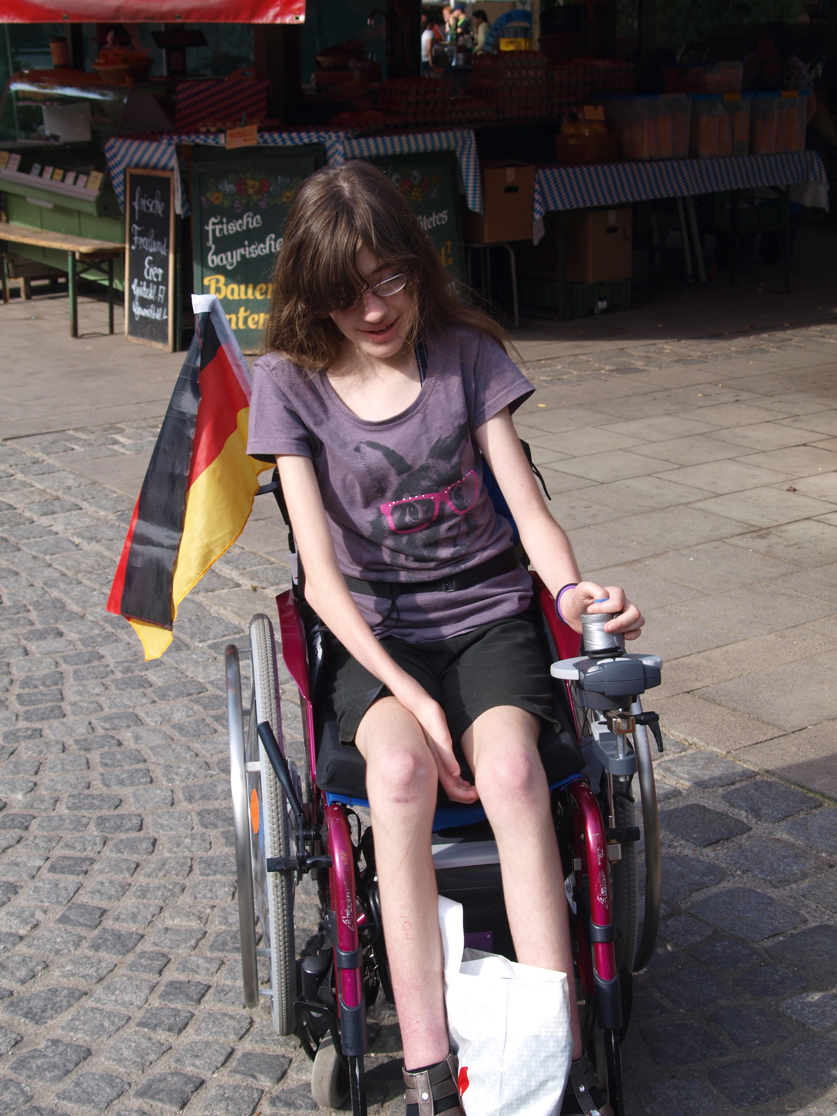 Jugendliche-Version (ca. 14 Jahre alte) von Laura fährt in einem Aktiv-Rollstuhl mit Motor-Antrieb durch eine Straße in München. Auf dem Fußbrett, zwischen den Füßen transportiert sie eine Tüte. Hinten am Rollstuhl hängt eine Deutschland-Fahne.