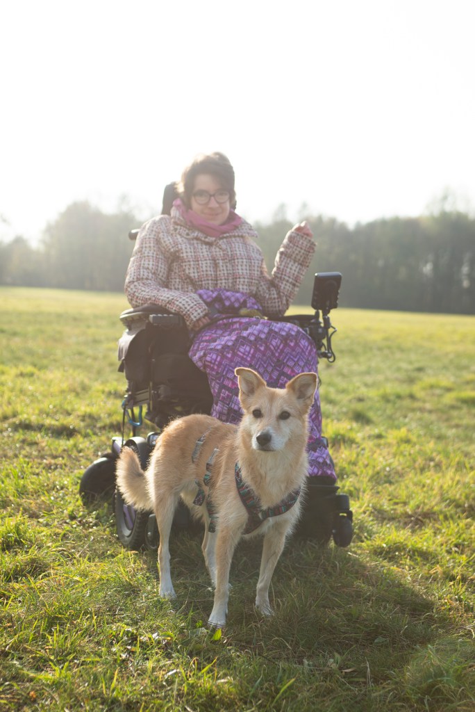 Laura steht mit dem Elektrorollstuhl auf einer grünen Wiese. Sie hat einen Fußsack mit einem Muster in der Farbe pink an und trägt eine karierte Jacke. Sie hat kurze Haare. Vor ihr steht ein kleiner Hund in beige. Er schaut aufmerksam, mit aufgestellten Ohren die Kamera. Das Bild ist im oberen Bereich sehr hell, was die Kulisse etwas magisch erscheinen lässt.
